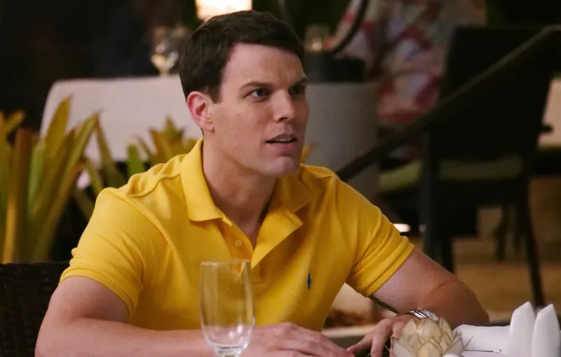 A man in a yellow polo shirt sits at a table in a restaurant, looking to the side. There are empty glasses and folded napkins on the table. The background is softly blurred, with plants and dim lighting.