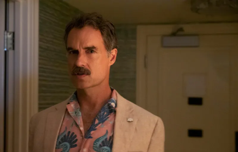 A man with a mustache and short hair stands indoors, wearing a light-colored blazer over a pink and blue patterned shirt. The background features a partially open door and textured wallpaper. The lighting is warm and dim.