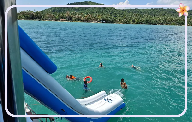 People swim in clear turquoise waters near a blue and white inflatable slide extending from a boat. Green hills and palm trees are visible in the background. A red circular float is being used by one of the swimmers.