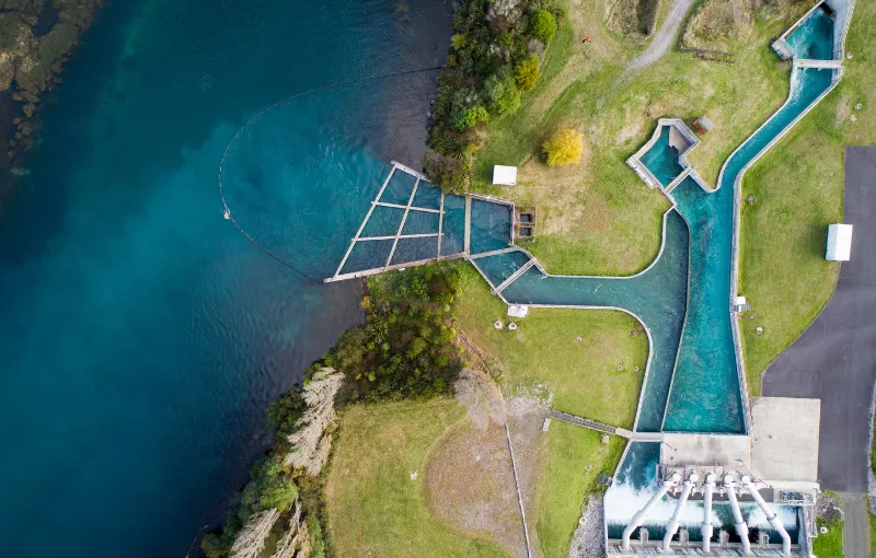 Aerial view of a blue water canal system with geometric structures, surrounded by green grass and small trees. The canal leads to a dam with water flowing through. A shoreline is visible on the left, with clear water showing natural formations.