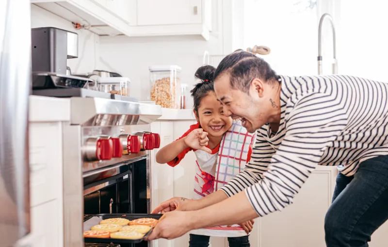 A man and a young girl are in a kitchen. The man, wearing a striped shirt, is putting a tray of food into the oven. The girl, wearing a red shirt and apron, smiles while giving a thumbs-up. They are surrounded by white cabinets.