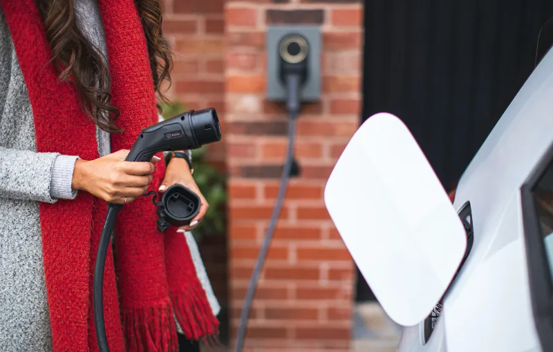 A person wearing a gray sweater and red scarf is holding a charging plug next to a white electric car. The car's charging port is open, and a wall-mounted charging station is visible on the brick wall in the background.