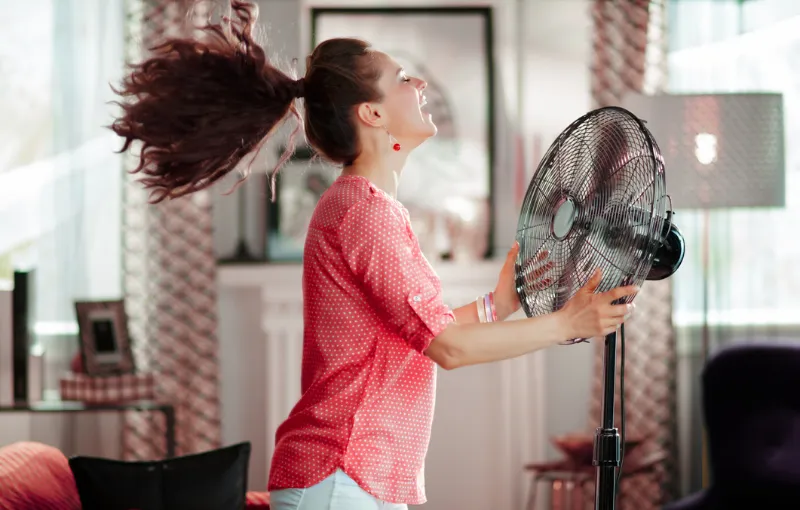 A woman wearing a red polka dot shirt stands indoors with her hair blown back by a large, oscillating fan. She appears joyful and is smiling, enjoying the breeze. The background includes patterned curtains and framed pictures.