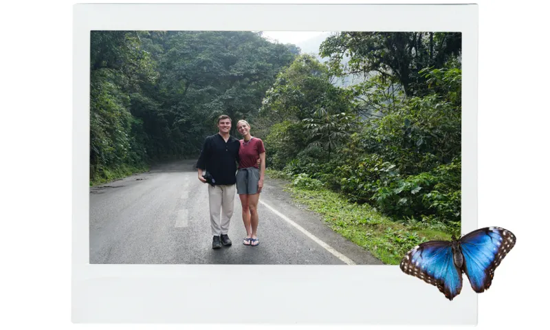 A man, Duncan, stands in a lush green section of jungle on tar sealed road. He is wearing light linen pants, hiking boots and a black linen top. Next to him is Liv a young woman with blonde hair wearing a red merino tee and gray hiking shorts and jandals, they have an arm around one another.