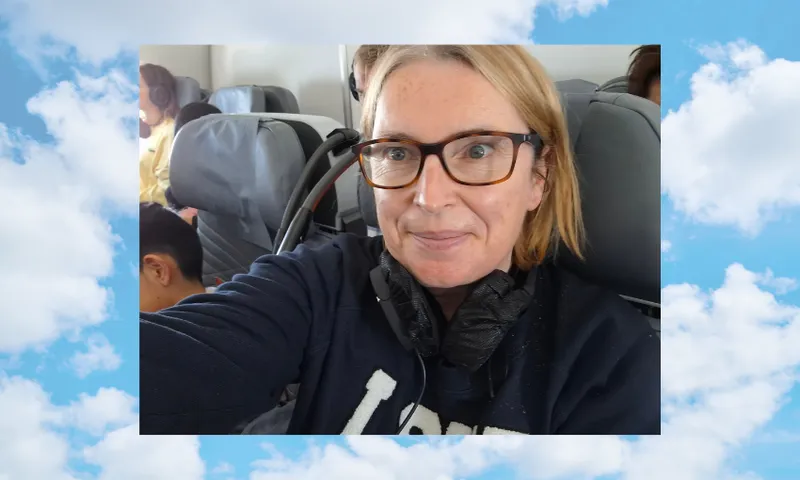 A woman with glasses and a headset, wearing a dark shirt, takes a selfie on an airplane. Passengers can be seen seated behind her. The image is framed with a background of clouds and blue sky.