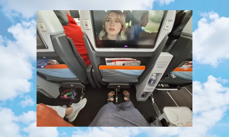 Airplane cabin interior showing two seat backs and entertainment screens. One screen displays a movie scene. A person’s legs are seen, wearing sandals on a footrest. Tray tables and personal items are visible. Blue sky and clouds frame the image.