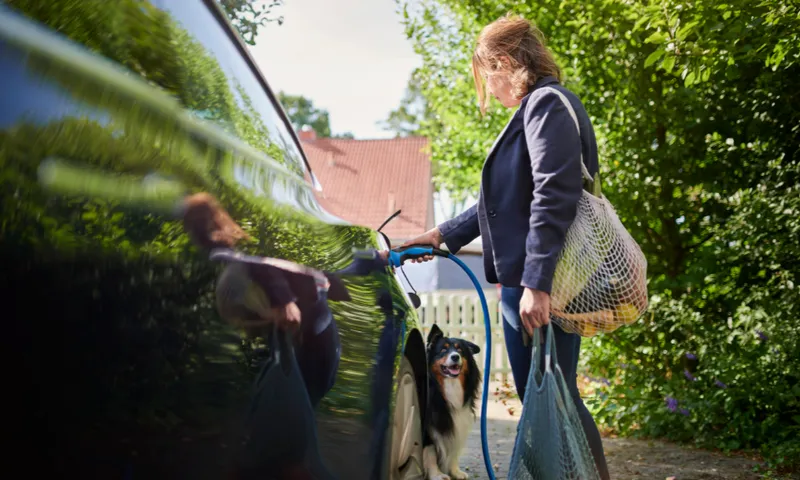 A woman is charging an electric car outdoors while holding a netted bag of groceries. A dog sits near her feet, looking up. The car's glossy surface reflects the greenery around them. The scene is bright and surrounded by trees.