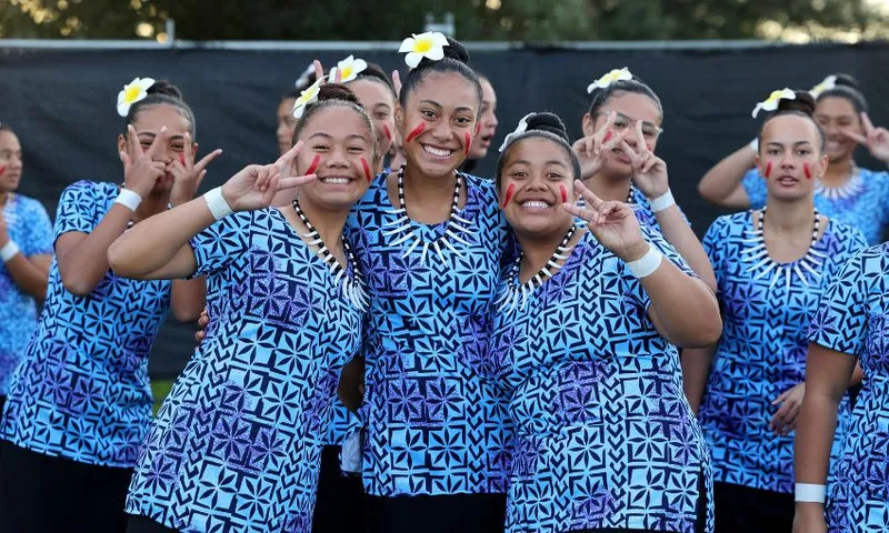 A group of smiling young women in matching blue patterned outfits pose together, some making peace signs. They wear jewellery and flowers in their hair, they are standing outdoors in front of a dark background.