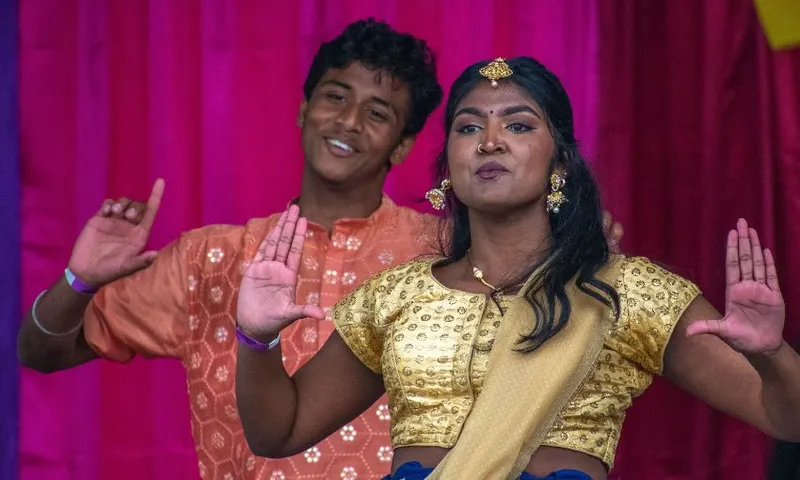 Two people in traditional attire perform a dance against a pink and red backdrop. The person in front wears a gold blouse and yellow sash, while the person behind wears an orange shirt. Both have raised hands and smile expressively.
