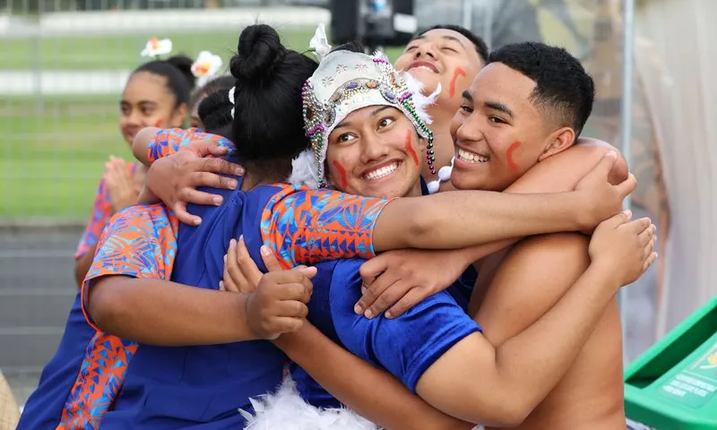A group of young people in colourful attire are joyfully hugging each other. They wear traditional clothing, with one person in a detailed headdress. Everyone is smiling, with visible face paint, showing a sense of celebration and togetherness.