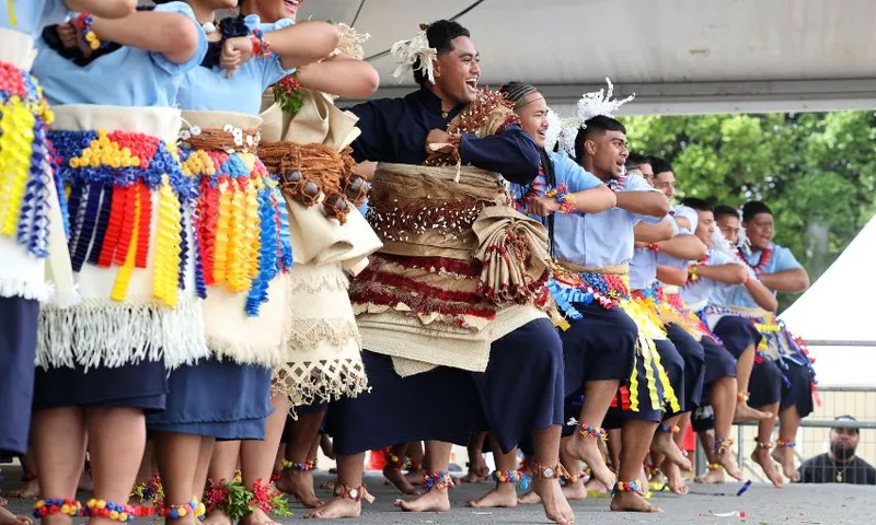 A group of people wearing colourful traditional Pacific Island attire performs a dance on stage. They are smiling, with festively decorated clothing, and adorned with flowers and woven accessories. Trees are visible in the background.