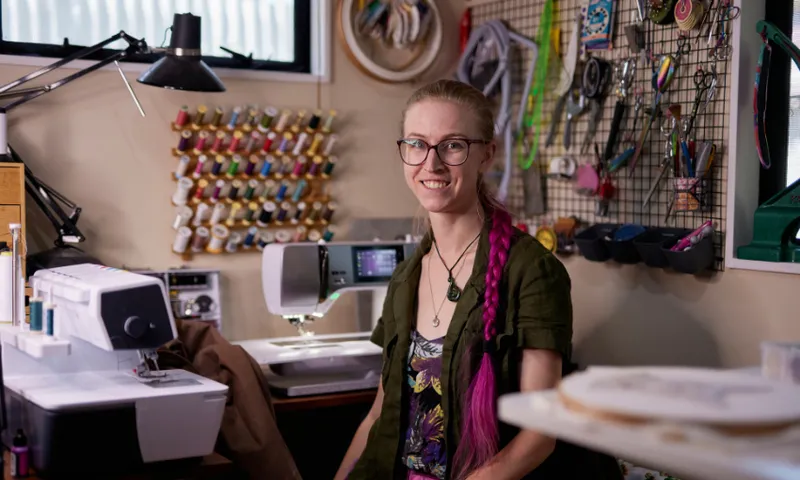 Kat Waters is wearing glasses and a long pink braid. She sits in a sewing room. Behind her are sewing supplies, spools of thread on a rack, and various tools on a pegboard. She is near a sewing machine, smiling, in a well-lit space.