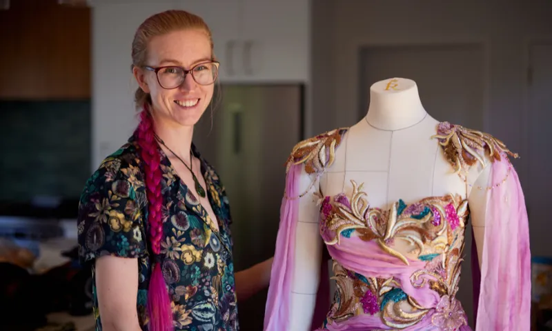 Kat Waters is shown with long pink hair and glasses. She smiles while standing next to a dress form. The dress form features an ornate costume with purple fabric and gold decorative details. The background includes a kitchen setting.
