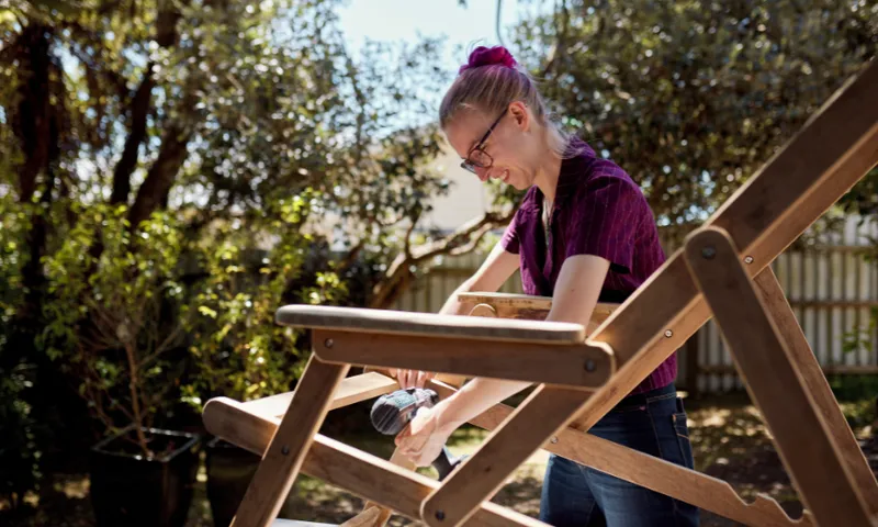 Kat Waters is wearing glasses and a purple shirt. She uses a small electric sander on a large wooden chair outdoors. Sunlight filters through trees in the background, creating a lively garden atmosphere.
