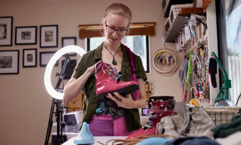 Kat Waters examines a bright pink boot she made in a room filled with crafting supplies. A ring light and various tools are visible. Framed pictures hang on the wall in the background.