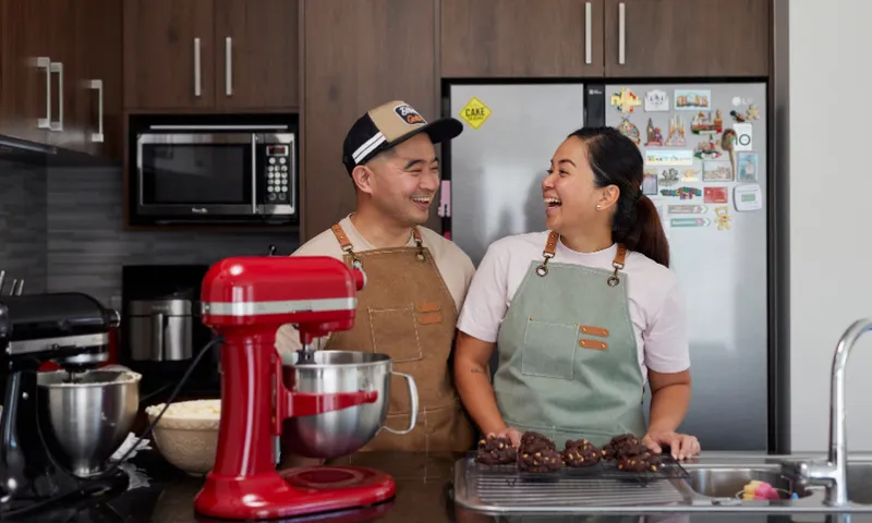 Jose Antonio and Nikki Villamil are seen both wearing aprons, smile at each other in a kitchen. They stand near a red stand mixer with bowls and chocolate treats on the counter. The kitchen has wooden cabinets and a fridge with magnets.