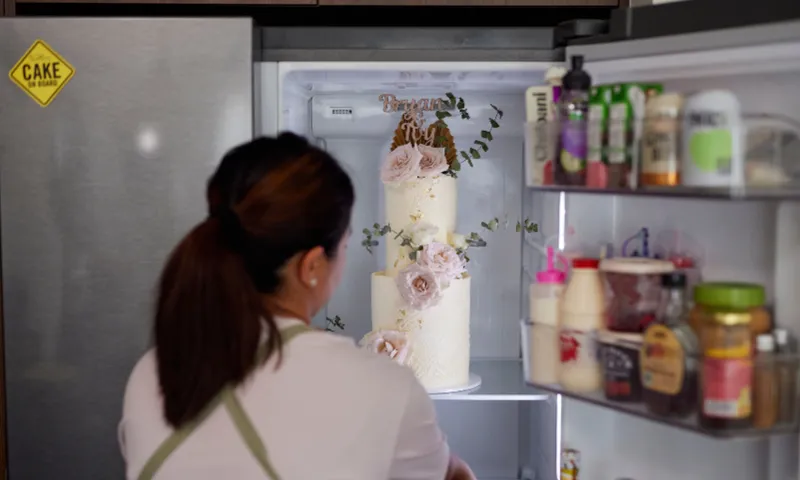 Nikki is seen from behind, looking at a tall, elegantly decorated cake inside an open fridge. The cake is adorned with pink flowers and greenery. The fridge door shelves hold various condiments and bottles. A "CAKE" sticker is visible on the fridge exterior.