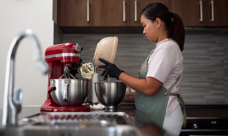 Nikki Villamil is wearing an apron and gloves and pours batter from a bowl into a mixing bowl of a red stand mixer in a modern kitchen. The kitchen has wooden cabinets and stainless steel appliances.