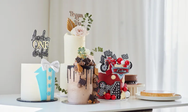 A display of four decorative cakes on a table. Each cake has unique themes: one with a bow tie, another with chocolate drips and treats, a floral one with golden accents, and a racing-themed cake. A plain tart is in the background.