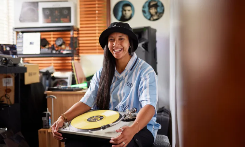 DJ Leemz is wearing a bucket hat and a striped shirt. She smiles while sitting in a room with music equipment. She holds a yellow vinyl record with a turntable. Decor includes music-themed items on the wall