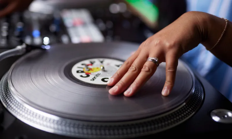 A close-up of DJ Leemz's hand expertly scratching a vinyl record on a turntable. The hand is adorned with a silver ring, and the turntable has a blurred colourful background, suggesting motion and activity in a lively setting.
