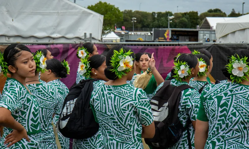 A group of women wearing green and white patterned dresses and floral headpieces gather outdoors. Some carry bags. Tents and trees are in the background. One woman in the centre gestures with two fingers raised in a peace sign.