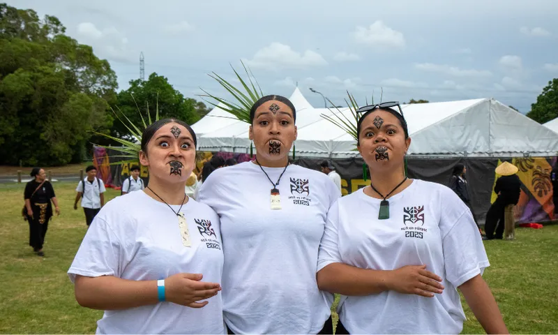 Three people dressed in white shirts and black headbands with ta moko face paint perform pukana with expressive looks. They wear pounamu and bone pendants and have leaves in their headbands. Tents and other people are visible in the grassy background.