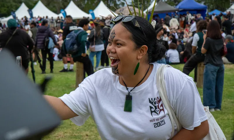 A woman with ta moko face paint and a carved pounamu pendant smiles, taking a photo at an outdoor cultural event. She wears a white shirt with "2025" printed on it. Tents and people are visible in the background.
