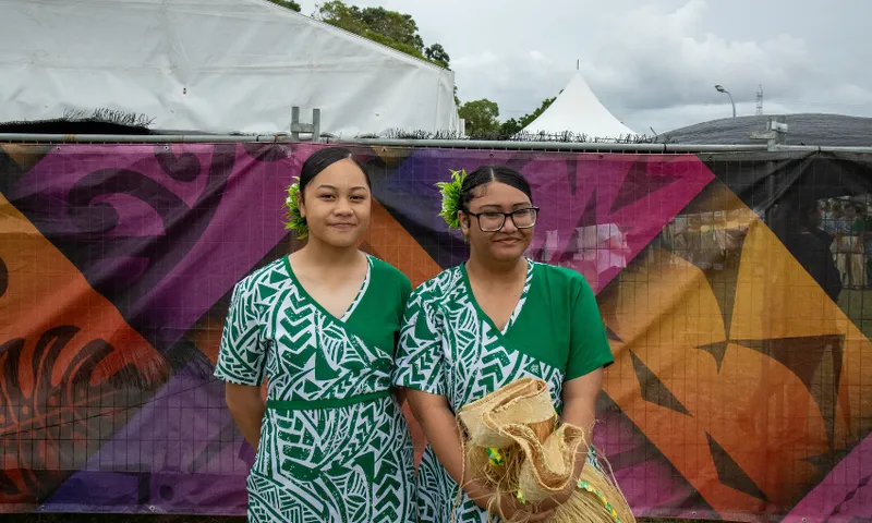 Two women in green and white patterned dresses stand in front of a colourful geometric backdrop. They both have flowers in their hair. One woman holds a woven item. A white tent and trees are visible in the background.