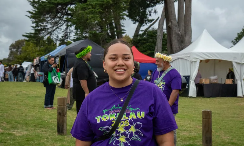 Luisa Ineleo is smiling at a cultural festival and wearing a purple shirt with "Tokelau" on it. Tents and other attendees in the background. Trees and a cloudy sky are visible.