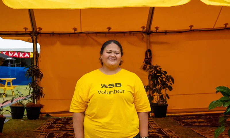 Lani Punatai in a yellow volunteer T-shirt stands smiling under a yellow tent. There are rugs on the ground and potted plants around. A bright blue structure and a red tent are visible in the background.