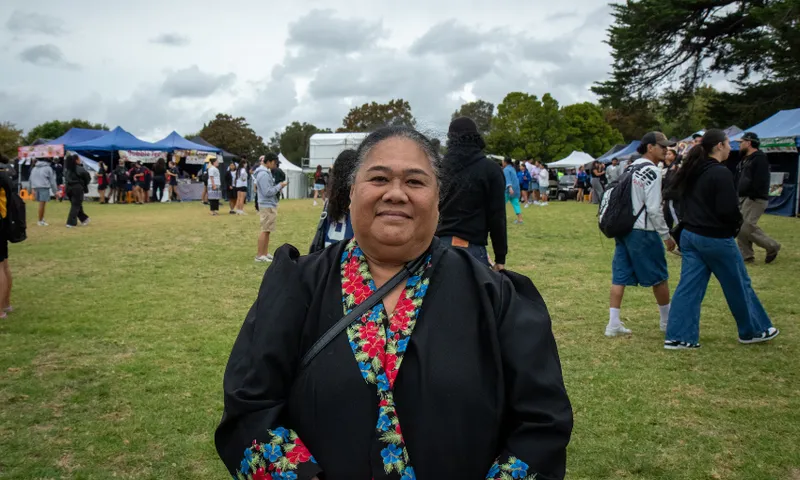 Mele Moliteugua is wearing a black outfit with colorful floral details and stands smiling in an outdoor festival setting. Tents and people are visible in the background on a grassy area under a cloudy sky.