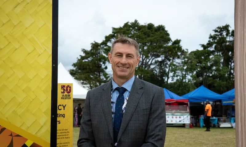 Steven Hargreaves in a suit stands smiling outdoors at an event. Behind him, there's a yellow sign with patterns, trees, blue canopies, and people in the background on a cloudy day.