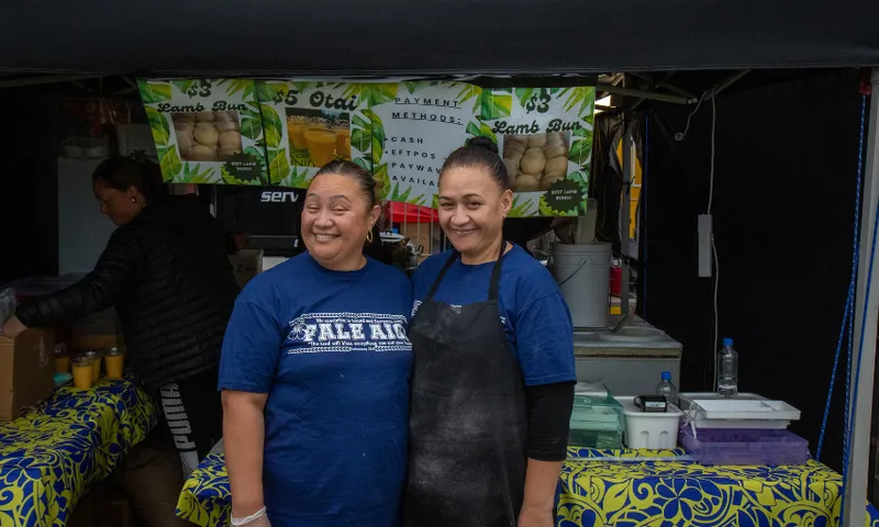 Anna and Josie Fruean stand smiling in front of a food stall with vibrant tablecloths and signs for lamb buns. They are wearing blue T-shirts and aprons. The stall is filled with various food items and equipment.