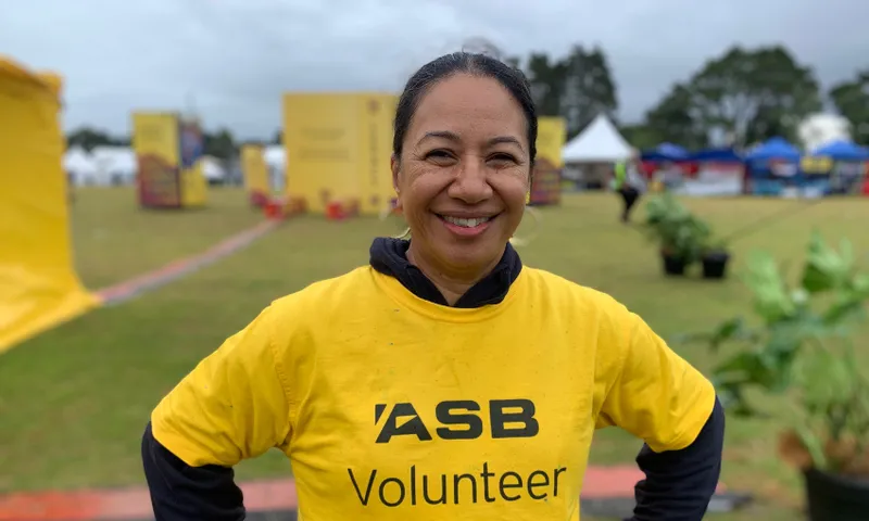 Teresa Tubu smiling and wearing a yellow "Volunteer" T-shirt stands outdoors at an event. The background features several tents and a grassy area under a cloudy sky.