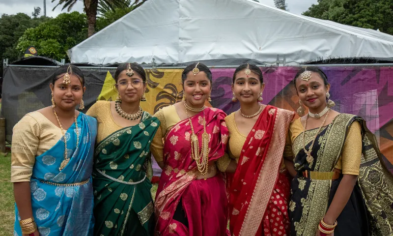 Five people stand side by side, smiling and wearing colorful traditional outfits with intricate patterns. They are dressed in shades of blue, green, red, and gold. A large tent and trees are visible in the background.