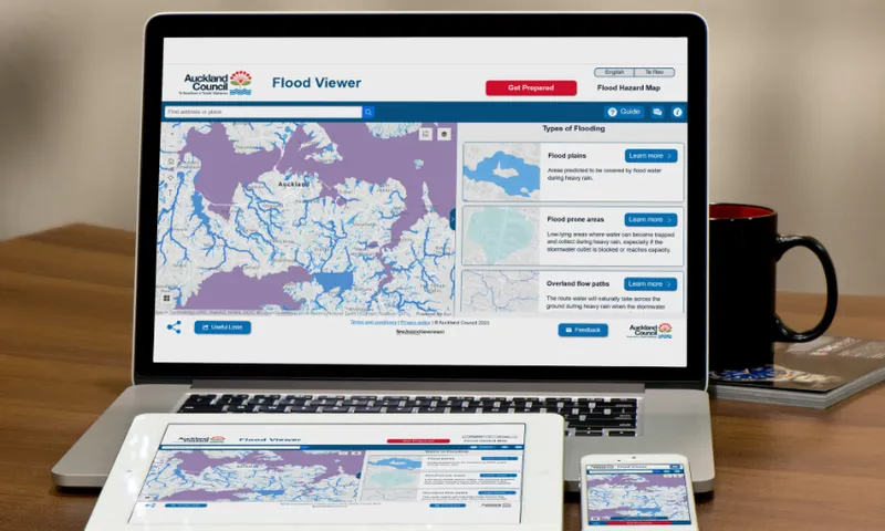 A laptop, tablet, and smartphone display a digital map with purple flood zones from the Auckland Council Flood Viewer website. Text and details about flood plains, flood zones, and overland flow paths are visible alongside the map. A red mug sits nearby.