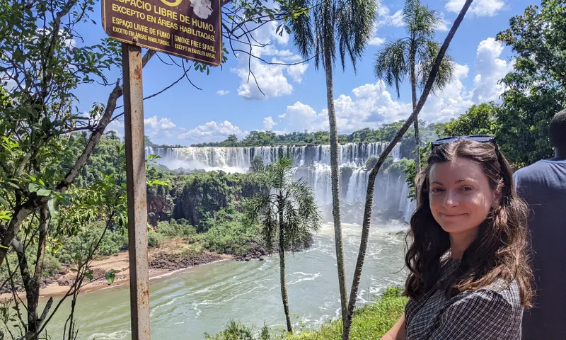 A woman stands in front of the Iguazu Falls, surrounded by lush greenery under a bright blue sky. A sign in Spanish nearby indicates a smoke-free area.