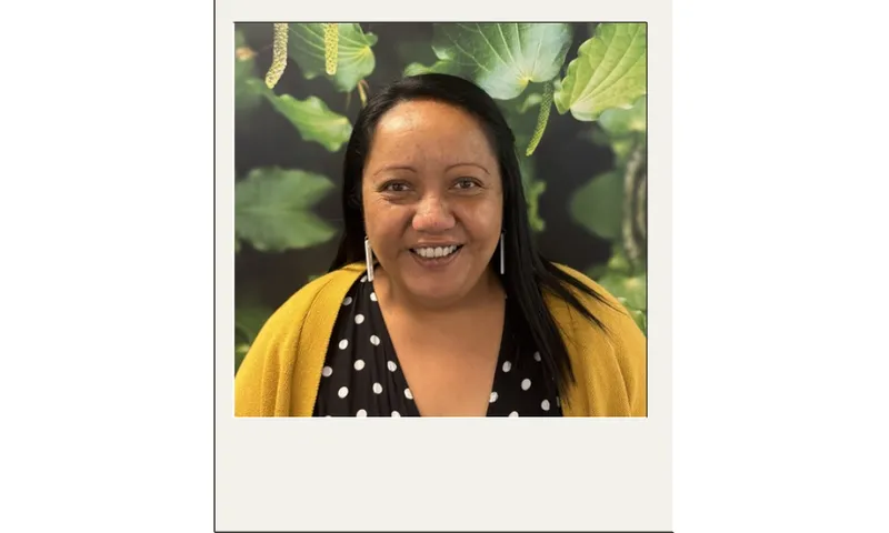 Priscilla Laita, a woman with long black hair smiles at the camera. She is wearing a black top with white polka dots and a mustard yellow cardigan. The background features large green kawakawa leaves and berries.