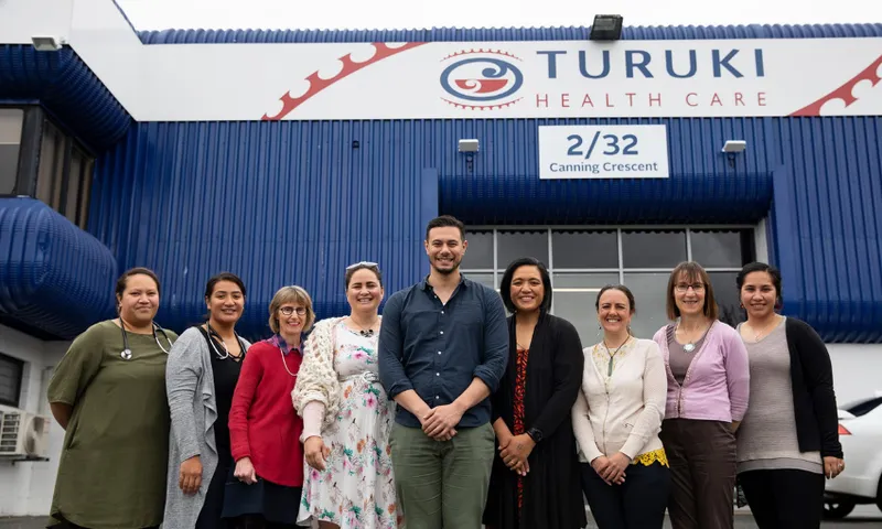 A group of people standing in front of Turuki Healthcare and smiling in front of a building with "Turuki Health Care" signage. The building is blue and white, located at 2/32 Canning Crescent. The group appears cheerful and casually dressed.