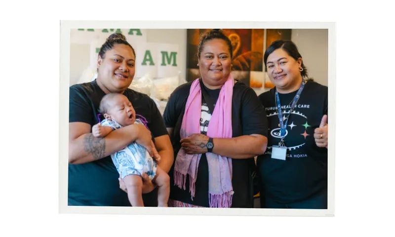 Three adults stand together indoors, smiling at the camera. One woman holds a baby wrapped in a light blue patterned blanket. The two others are wearing black T-shirts, and one is giving a thumbs up.