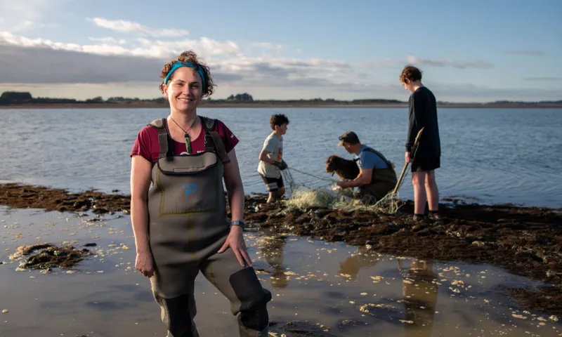 Jane Kitson in waders stands smiling by the shore, while three people and a dog work with nets in shallow water behind her under a blue sky.