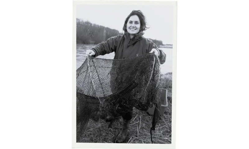 A smiling researcher, Jane Kitson, stands outdoors by a river, holding up a large fishing net. The scene appears to be during colder weather, as they are wearing a jacket. The image is in black and white.