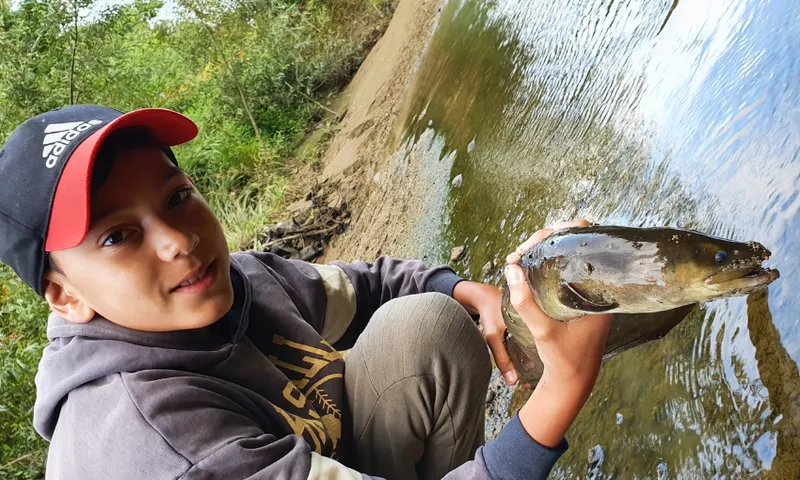 A boy wearing a red and black cap and grey hoodie kneels by a riverside, smiling as he holds a large tuna (freshwater eel) with both hands near the water’s edge. The background shows green foliage and a sandy bank.