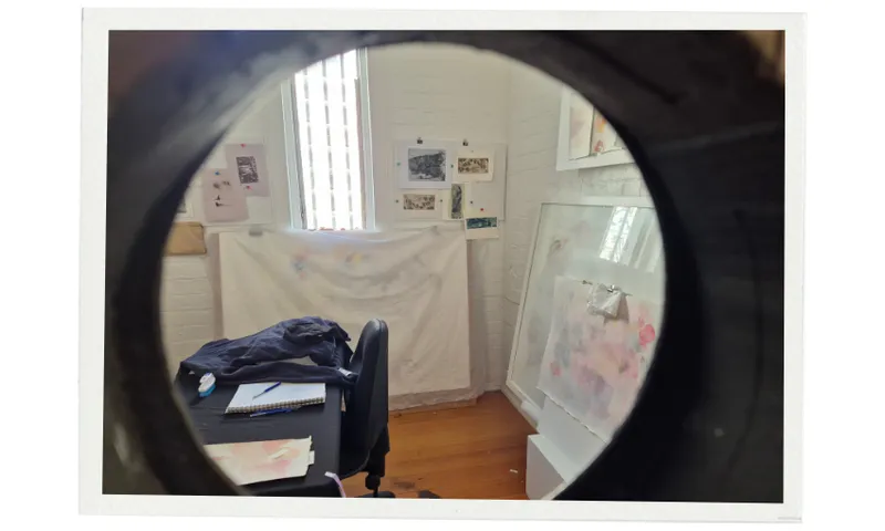 A view through a door's peep hole reveals a small art studio with sketches and art pinned to white walls, a table with a chair, a jacket, and a notebook, with natural light streaming through the windows.