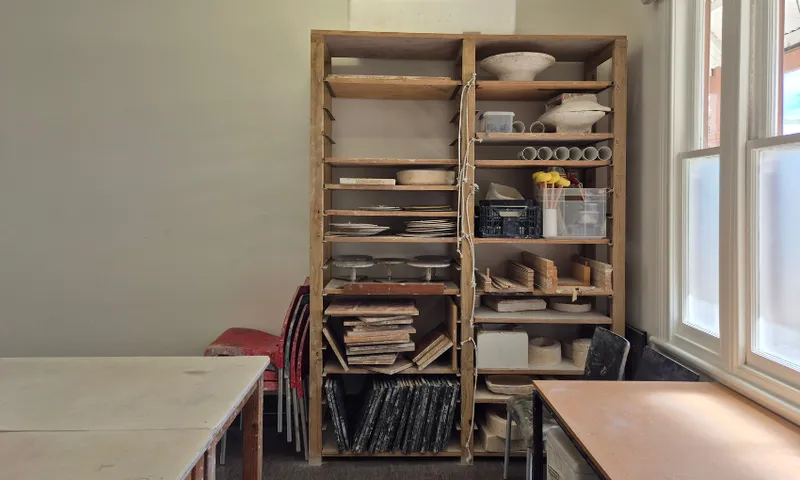 A wooden shelving unit holds pottery supplies, tools, and ceramic pieces in a well-lit room with large windows, tables, and stacked red chairs nearby.