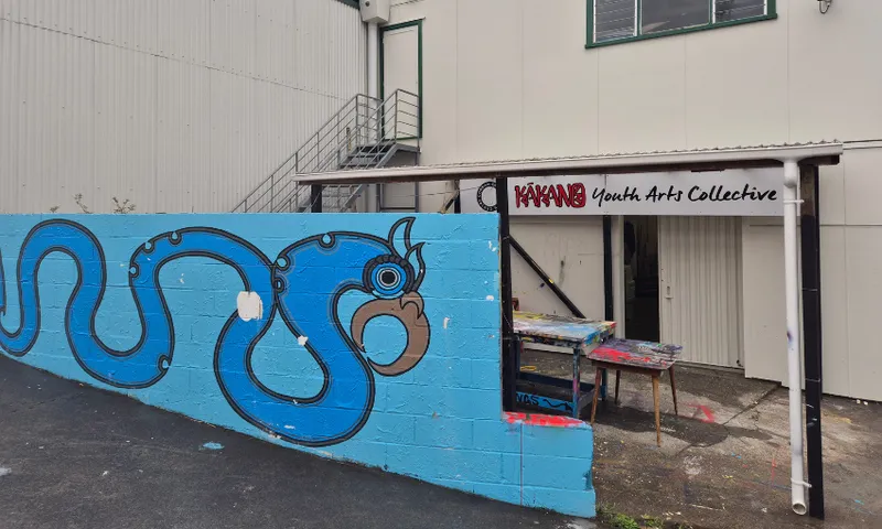 A blue mural of a stylised serpent or dragon decorates a low wall outside a building labeled "Kākano Youth Arts Collective." Colourful, paint-splattered tables are set up under the building’s awning.