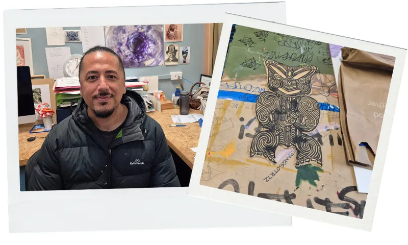 A man sits at a desk in a cluttered art studio with various papers and artwork behind him. Next to him is a detailed drawing of a Māori design laid out on a marked and used workspace.