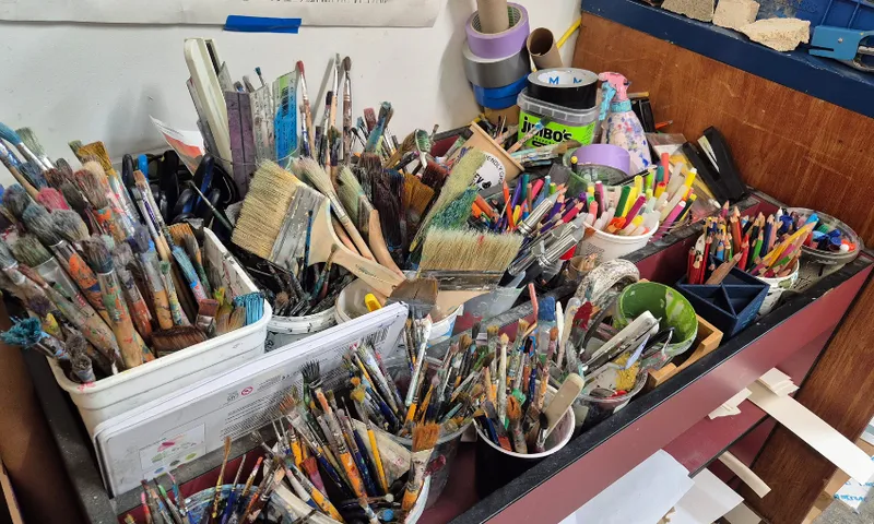 Assorted paintbrushes, markers, and art supplies are organized in various containers on a cluttered shelf in an art studio or classroom.