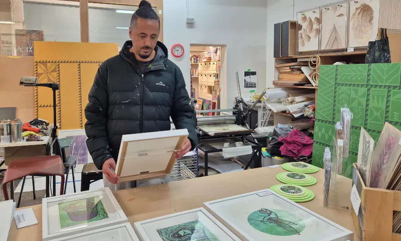 A person in a black jacket stands in an art studio, holding a framed artwork. Several framed artworks, prints, and art materials are displayed on the tables and walls around him.